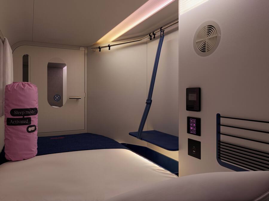 A modern hotel room with a bed, a small desk, a chair, a window, and a control panel.