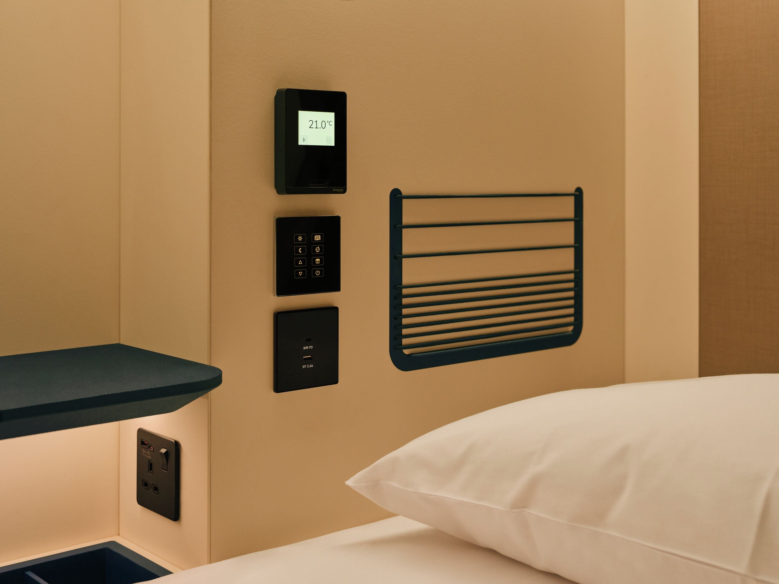 Ein Hotelzimmer mit Bedienfeld, das eine Temperatur von 21,0 °C anzeigt, einer Steckdose und einem schwarzen Handtuchhalter.