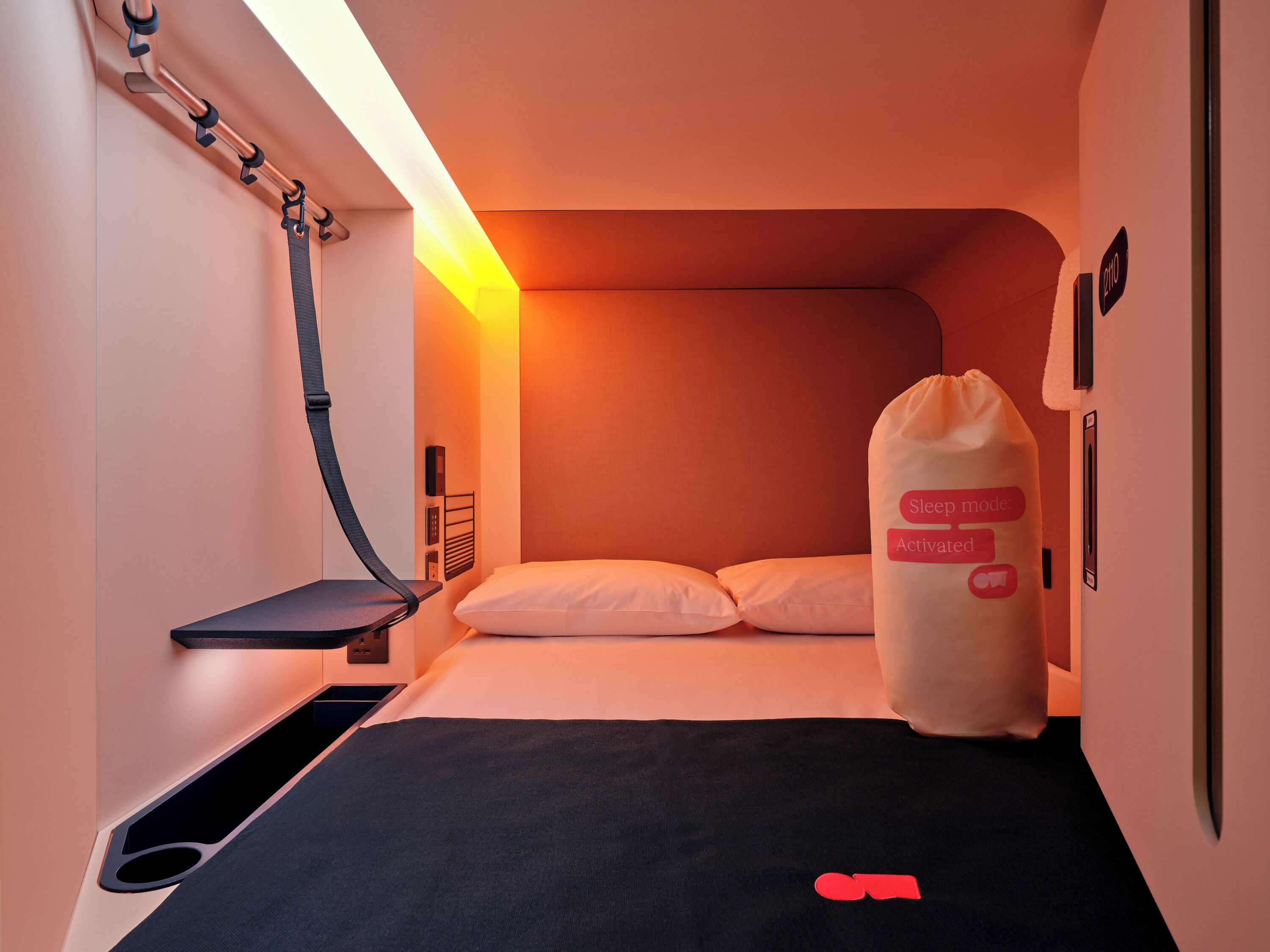 Ett kompakt hotellrum med en säng, ett sängbord, en kudde, en sänglampa och en väggmonterad kontrollpanel.