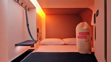 Ett kompakt hotellrum med en säng, ett sängbord, en kudde, en sänglampa och en väggmonterad kontrollpanel.