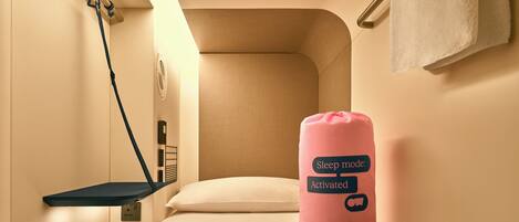 A compact room with a bed, a bedside table, a wall-mounted shelf, and a pink sleep mode indicator.