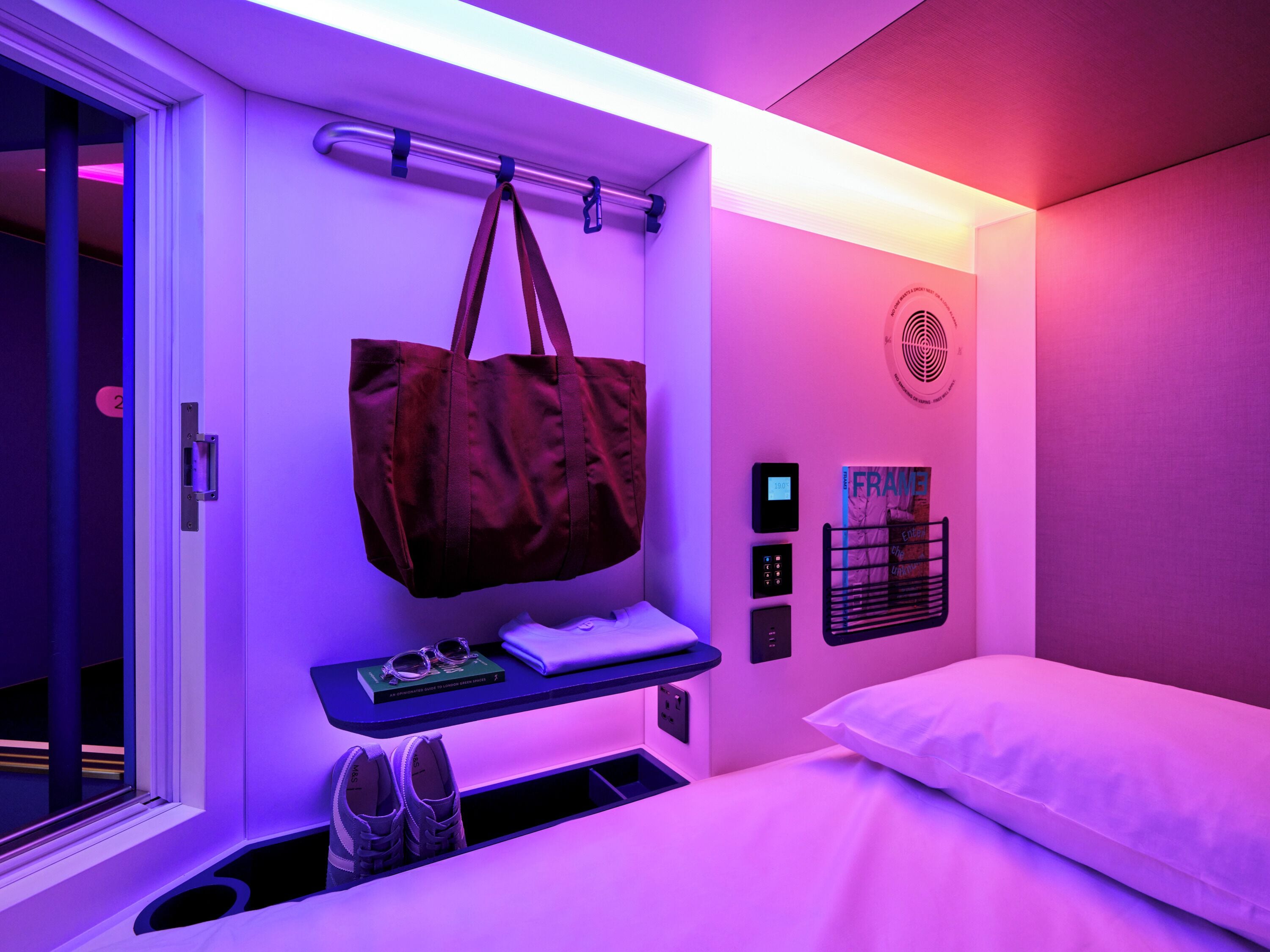 A hotel room with a bed, a bedside table with a laptop and glasses, a hanging bag, a shoe rack, and a wall-mounted control panel.