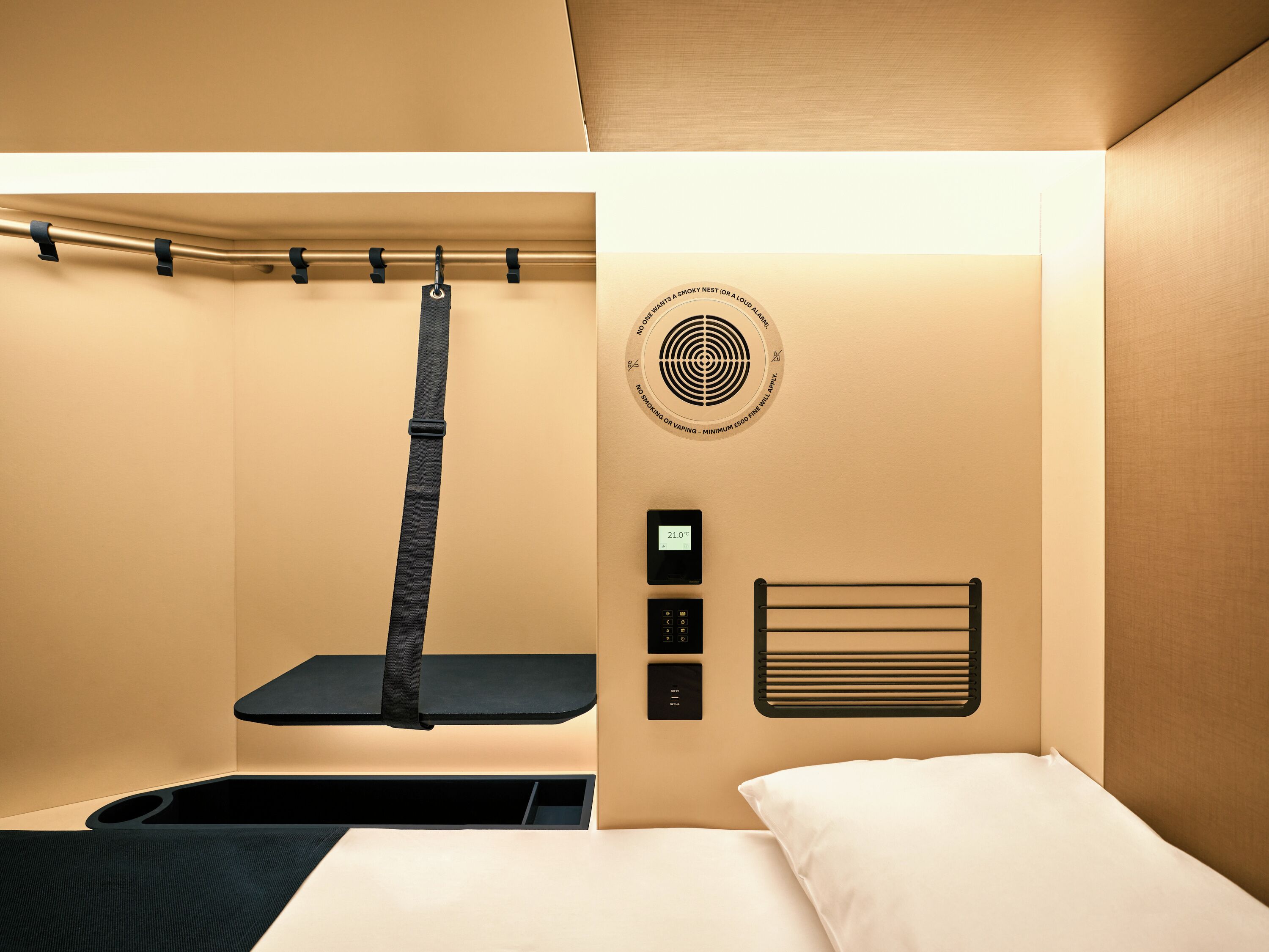 A modern hotel room with a bed, a small table, a wall-mounted heater, and a smoke detector.