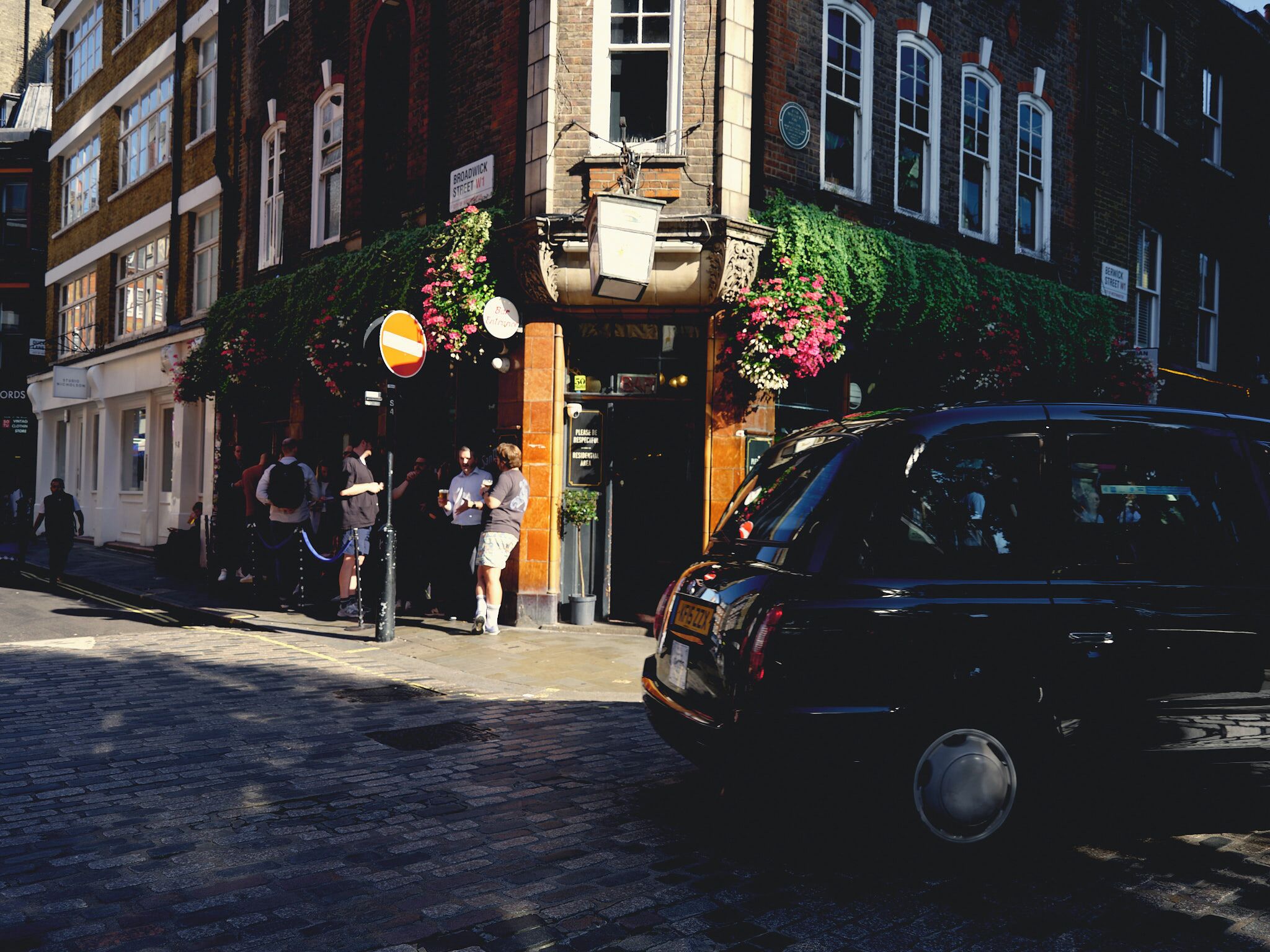 A street scene with a building featuring a brick facade, a sign reading 'BROADWICK STREET,' and a black car parked on the street.