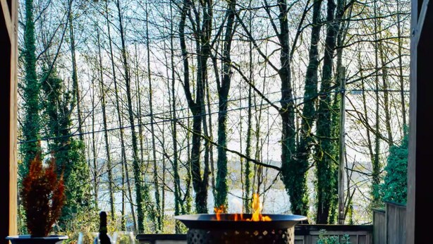 An outdoor patio with a fire pit, wooden tables, and chairs, surrounded by trees.