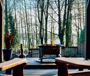 An outdoor patio with a fire pit, wooden tables, and chairs, surrounded by trees. - Cozy King Suite | Fire Pit | Near Lonsdale Quay - Pet Friendly (North Vancouver)