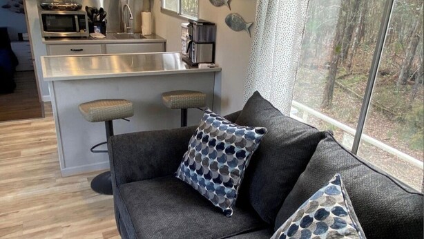 A modern living area with a grey sofa, patterned cushions, a kitchen with a microwave, and a large window offering a view of the outdoors.