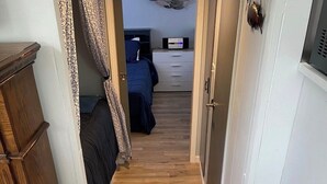 A narrow hallway with wooden flooring leading to a bedroom with a bed and a dresser.