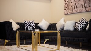Un sofá negro con cojines estampados, una mesa de centro con marco dorado y una alfombra blanca y esponjosa.