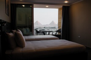Habitación de hotel con una cama y dos sillas frente a un balcón con vistas a pirámides.