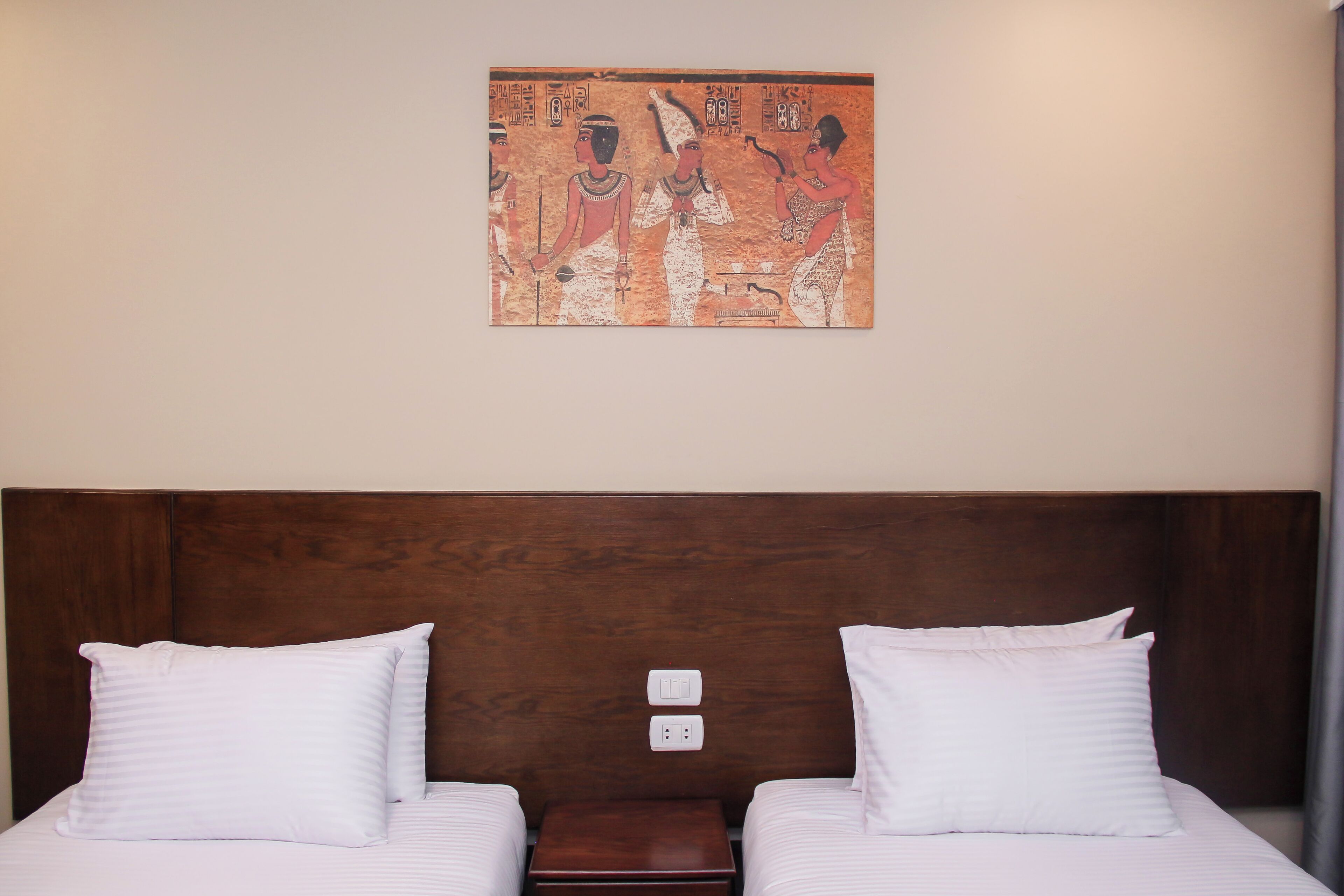 Ein Hotelzimmer mit zwei Betten, einem Holzkopfende, einem kleinen Nachttisch aus Holz und einem an der Wand hängenden Gemälde.