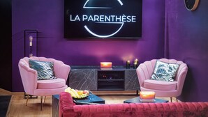 Un salon moderne avec un mur violet, un grand téléviseur à écran plat, deux fauteuils roses, un canapé rouge et un meuble de rangement noir pour les appareils électroniques.