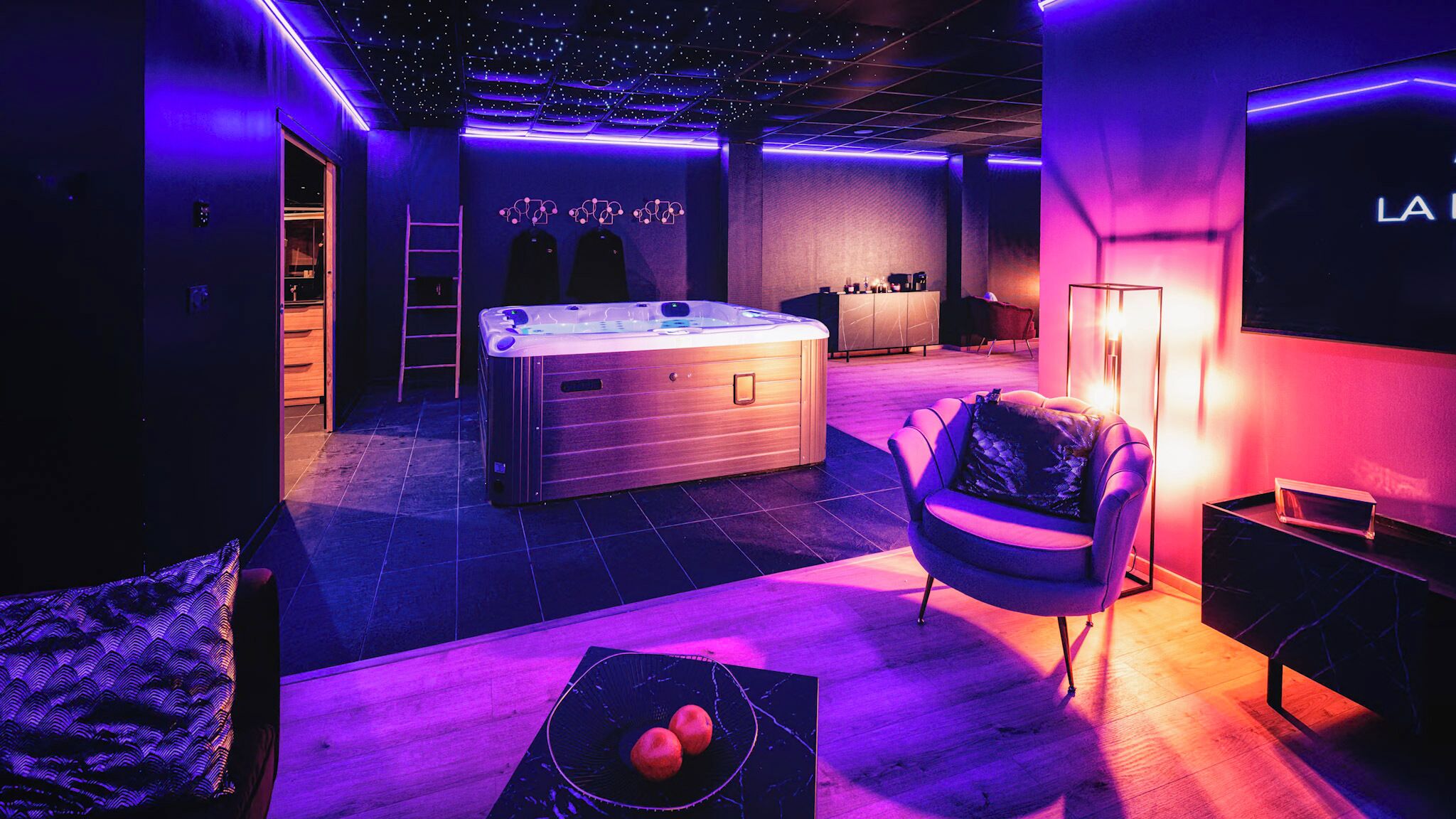 A modern, dimly lit room with a large hot tub, a cozy seating area, and a bar.