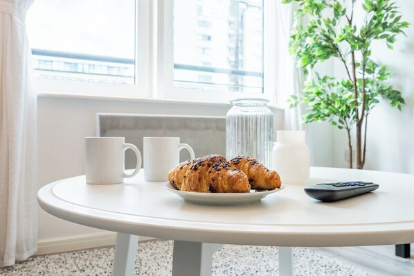 A round white table with a plate of croissants, two white mugs, a glass jar, and a remote control.