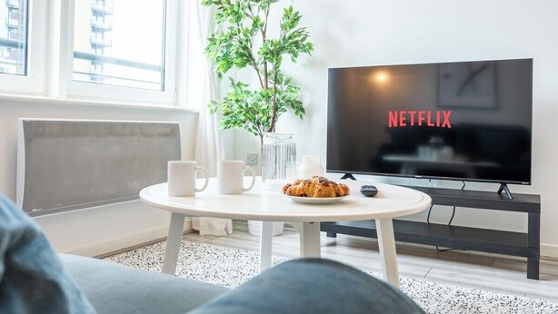 Un soggiorno moderno con una grande TV a schermo piatto che mostra "NETFLIX", un tavolo rotondo bianco con un piatto di cibo e due tazze bianche, una pianta in vaso e un divano grigio.