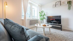 Un soggiorno moderno con un divano grigio, un tavolino rotondo, una TV che mostra 'NETFLIX', una pianta e una finestra con tende.