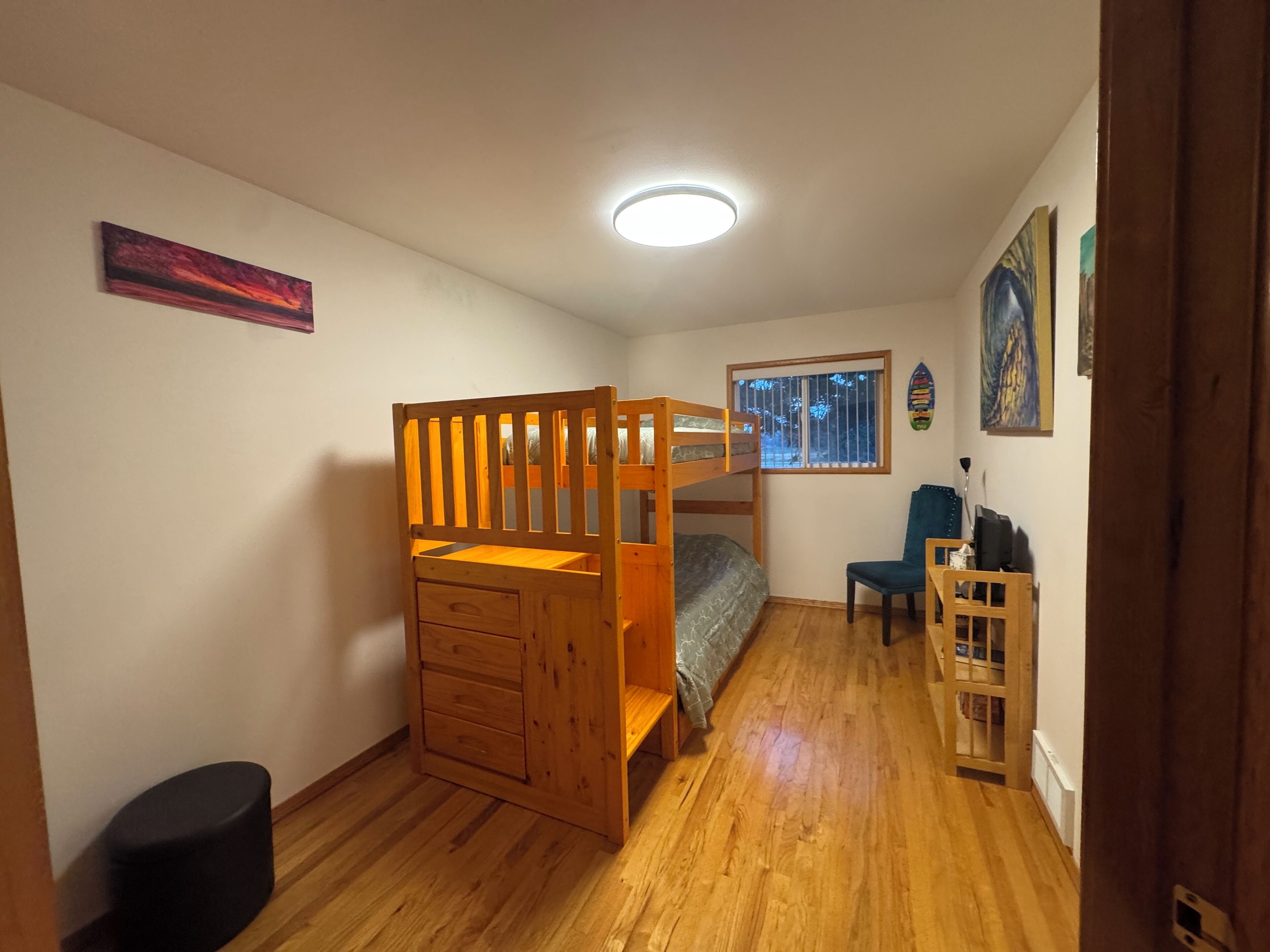A room with a bunk bed, a desk, a chair, a small stool, and a window with bars.