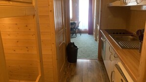 Een smalle houten gang die leidt naar een keuken met een spoelbak, fornuis en kasten. Een prullenbak staat op de vloer.