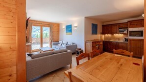 Interno di una baita in legno con zona pranzo, angolo cottura e divano accanto a una porta scorrevole in vetro.