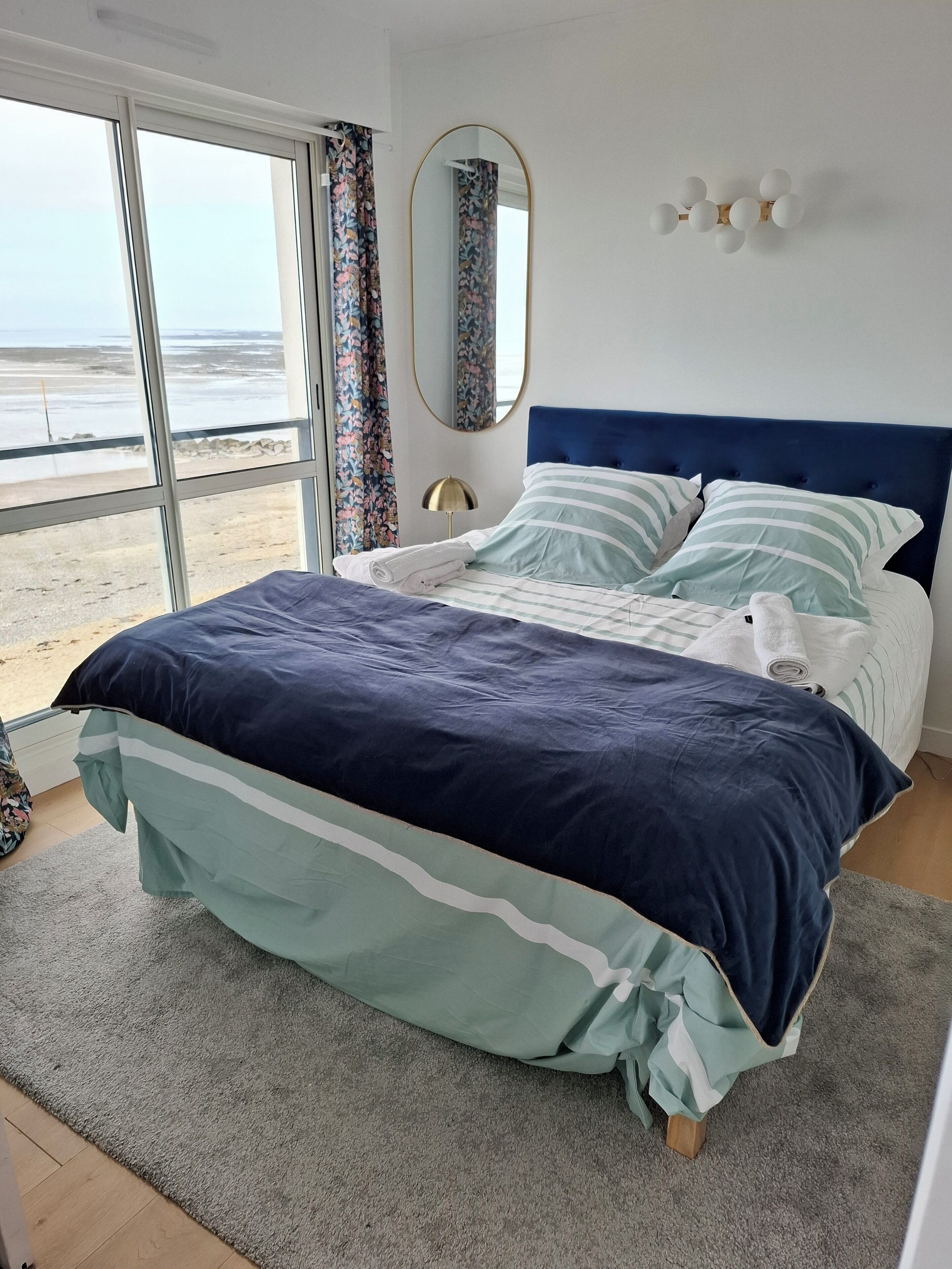 A bedroom with a large bed, a view of the beach, a mirror, and a wall-mounted light fixture.