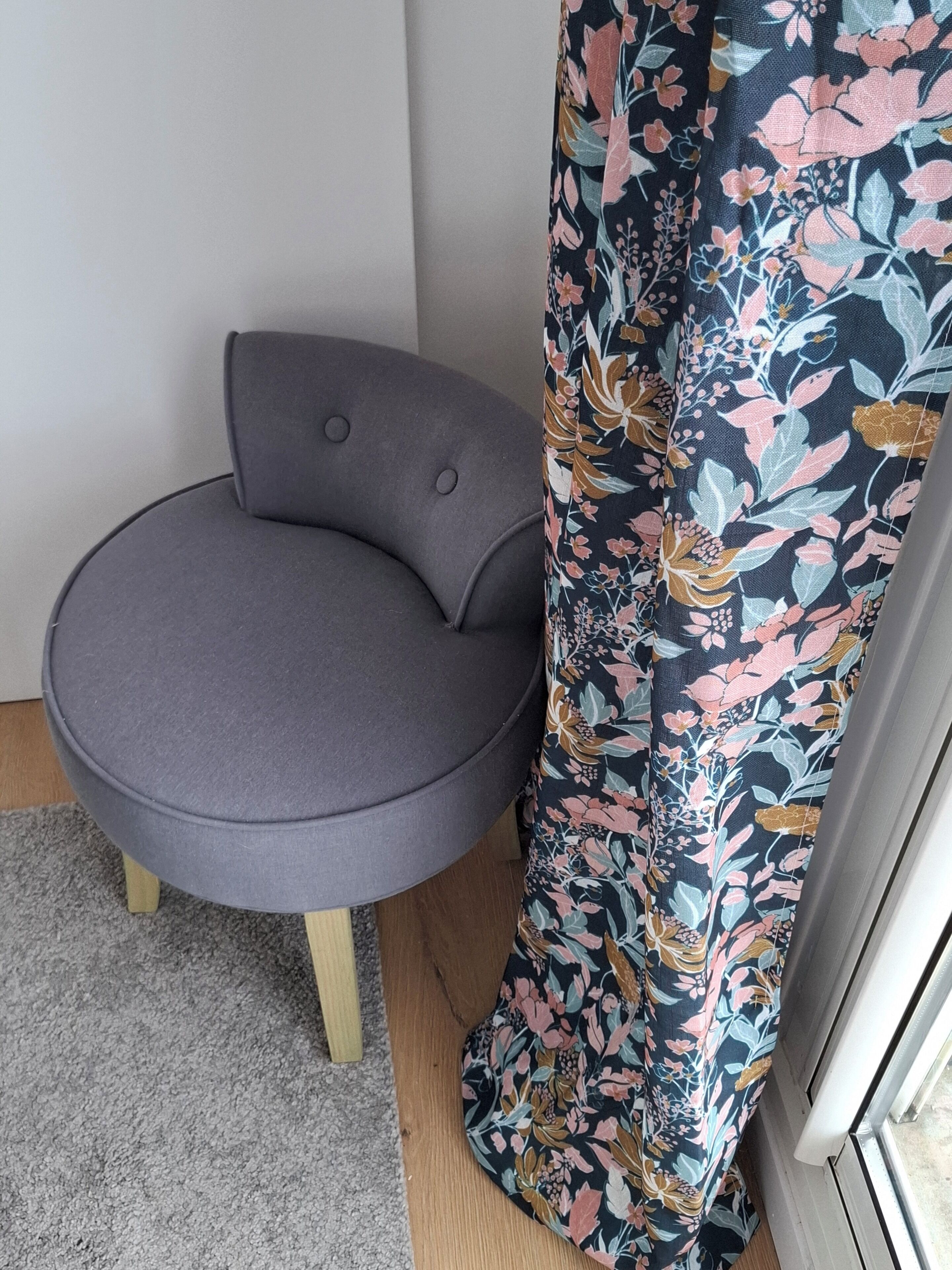 A corner of a room with a grey upholstered chair, a floral-patterned curtain, and a grey carpet.