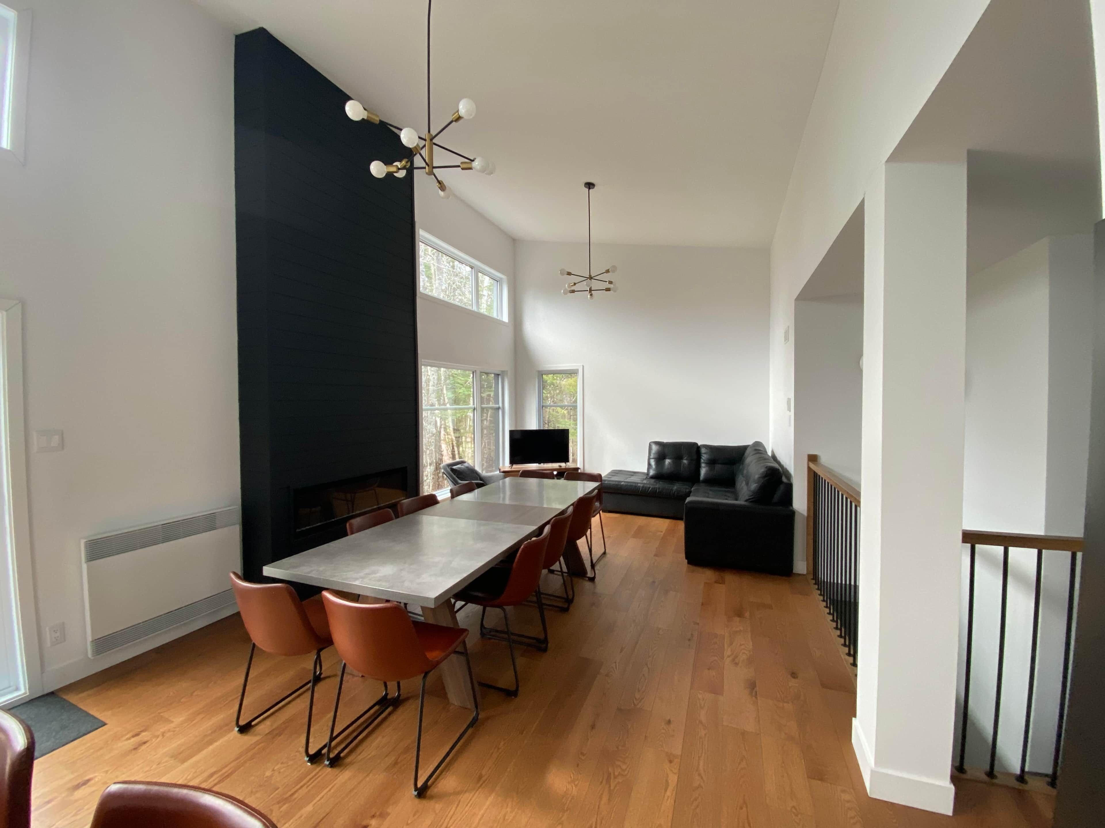 A spacious living area with a long dining table, multiple chairs, a black leather sofa, and a modern chandelier.