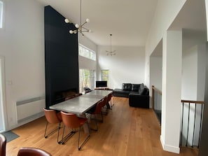 Een ruime woonkamer met een lange eettafel, meerdere stoelen, een zwarte leren bank en een moderne kroonluchter.