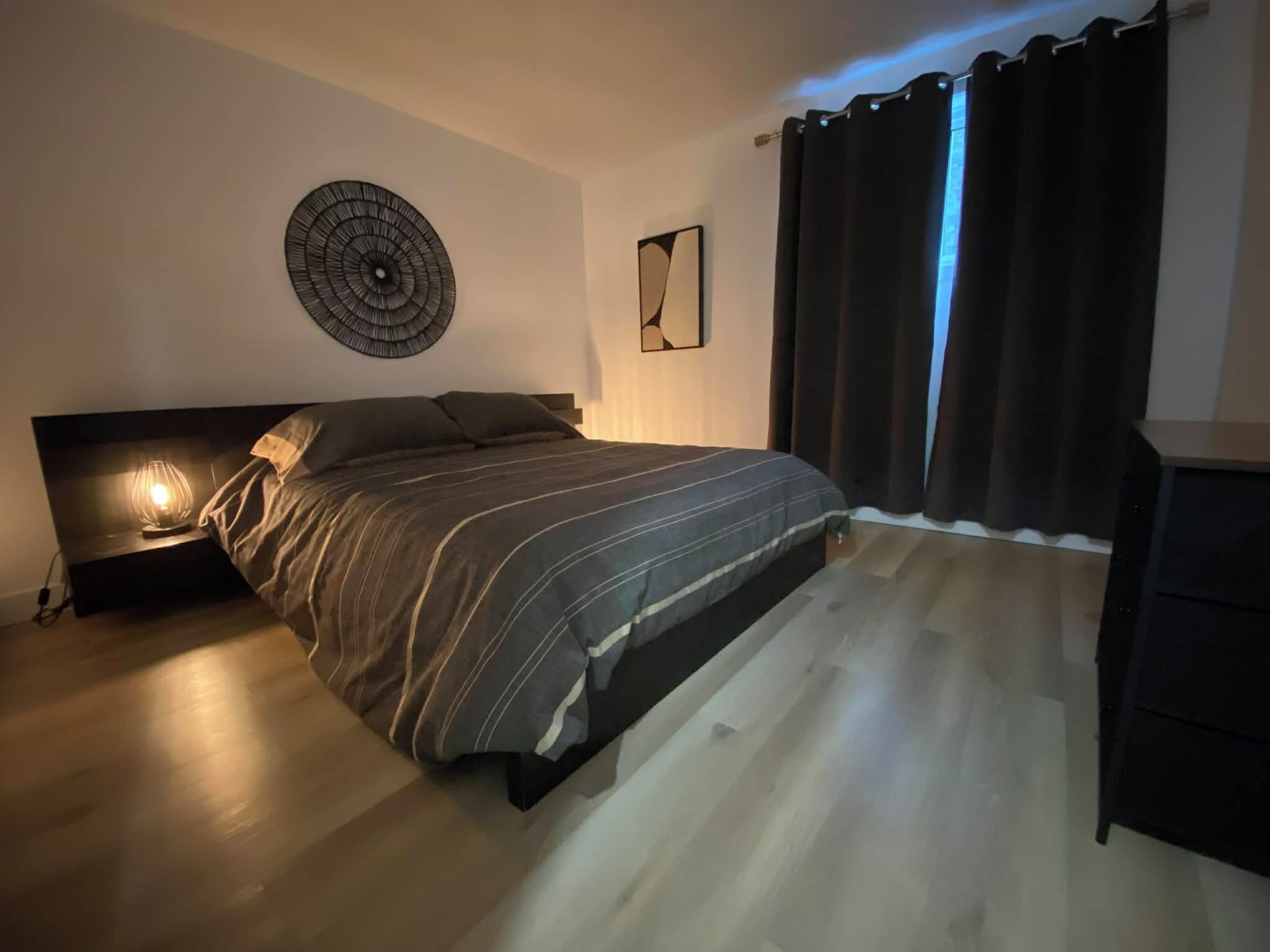 Un dormitorio con cama, lámpara de noche, una obra de arte en la pared y una ventana con cortinas.