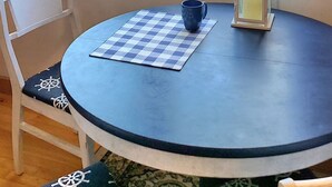 A room with a round table, a chair, a window with a view of a body of water and buildings, and a small lantern on the table. - Upper River House Waterfront Getaway on Ahnapee River with Dock Boat Slip Wifi (Algoma)