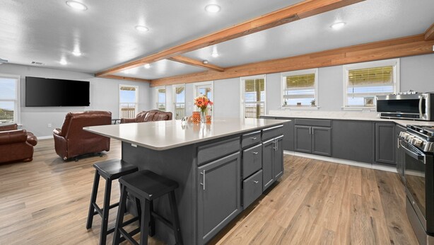 A modern kitchen with a large island, dark cabinets, wooden beams, and a flat-screen TV.
