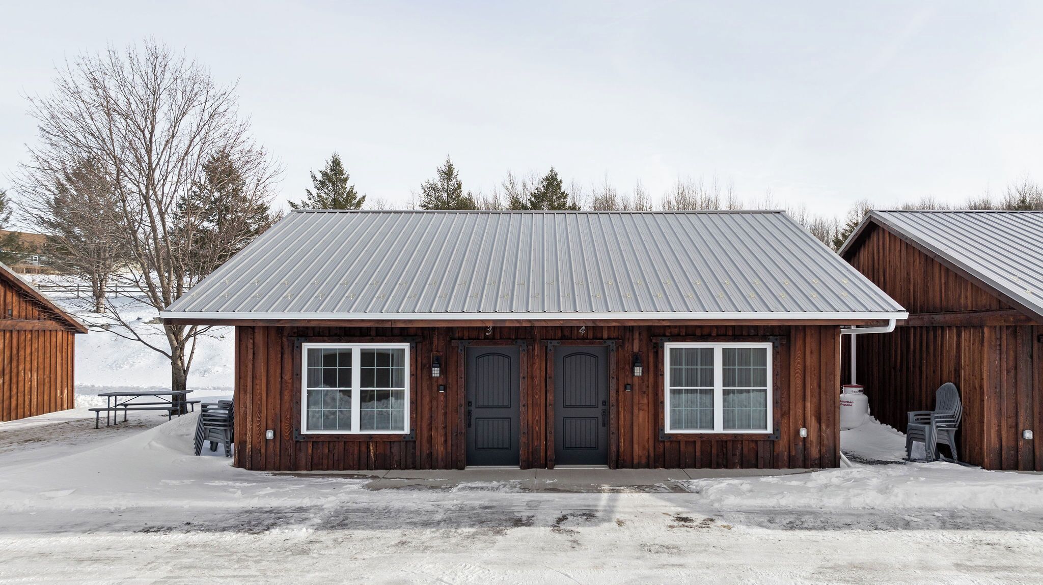 Un bâtiment en bois avec un toit en métal, deux portes d’entrée et un banc à l’extérieur.