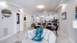 A modern living room with a round dining table, a sofa with decorative pillows, and a kitchen in the background.