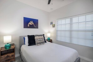 Une chambre à coucher avec un lit, deux tables de chevet avec des lampes, une fenêtre avec des stores et un tableau accroché au mur.