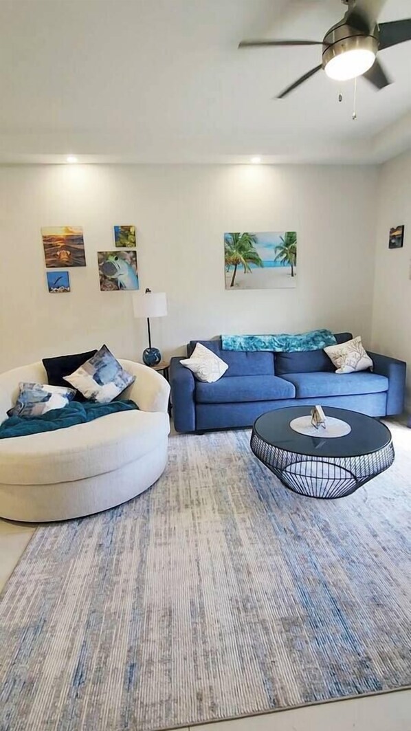Un salon avec un canapé blanc, un canapé bleu, une table basse ronde et un ventilateur de plafond.