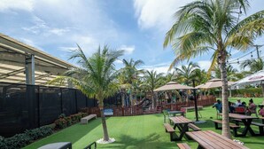 An outdoor patio area with artificial grass, wooden picnic tables, and palm trees.