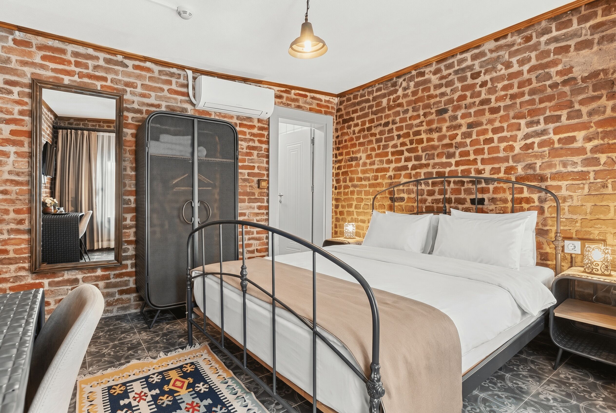 Una habitación con pared de ladrillo, cama con estructura metálica, armario, espejo y lámpara colgante.