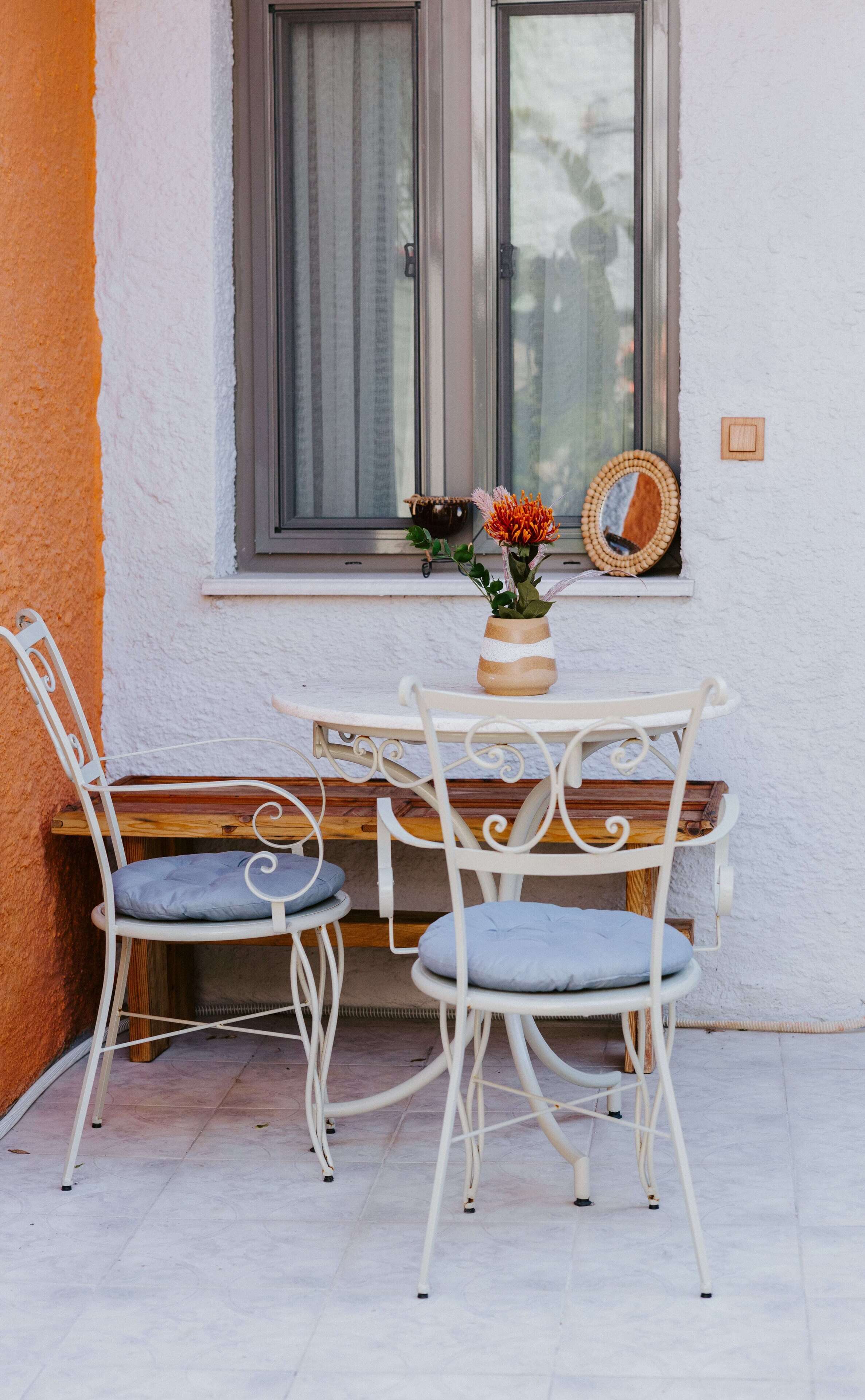 A small outdoor seating area with a table and two chairs, a window with curtains, and a vase with flowers.