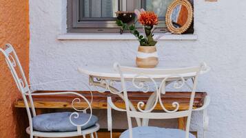 A small outdoor seating area with a table and two chairs, a window with curtains, and a vase with flowers.