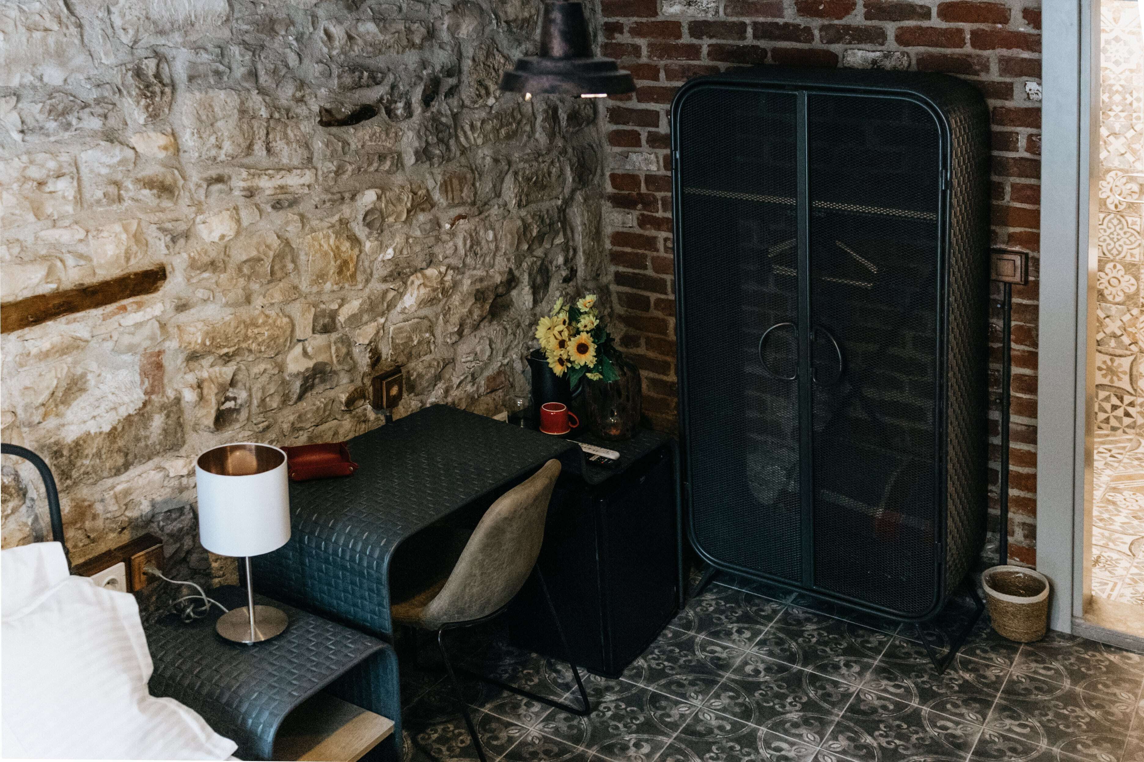 A rustic room with exposed brick walls, a metal cabinet, a small table with a lamp, and a chair.