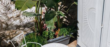Un patio con dos mesas redondas y cuatro sillas de metal, rodeado de plantas en macetas y un muro de piedra.