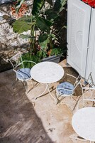 Um pátio com duas mesas redondas e quatro cadeiras de metal, cercado por vasos de plantas e um muro de pedra.