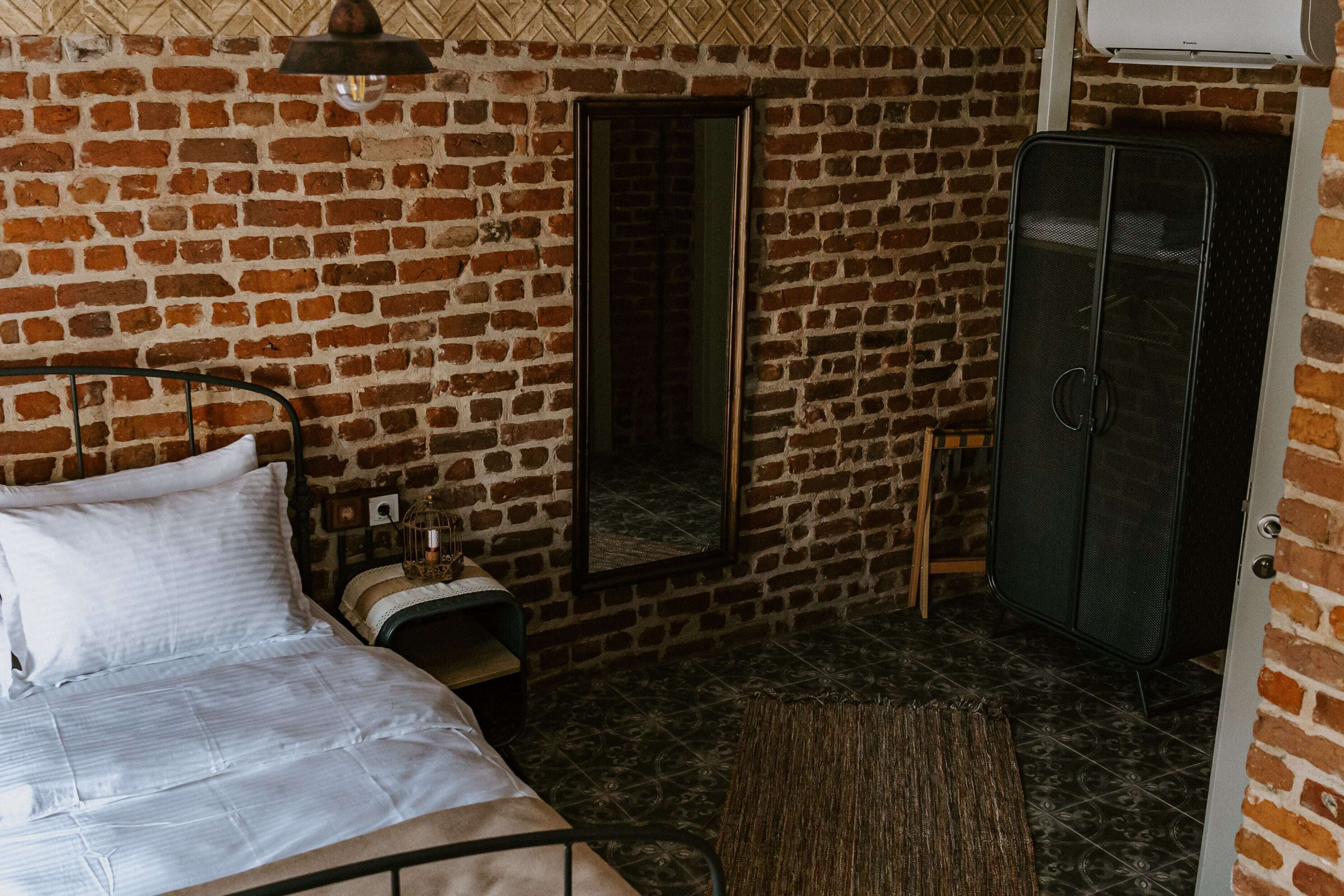 Une pièce avec un mur en briques, un lit avec une literie blanche, un miroir, une armoire et une petite table avec une lampe.