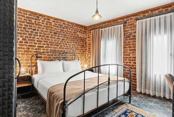 Un dormitorio con pared de ladrillo, estructura de cama metálica, mesita de noche con lámpara y ventana con cortinas.