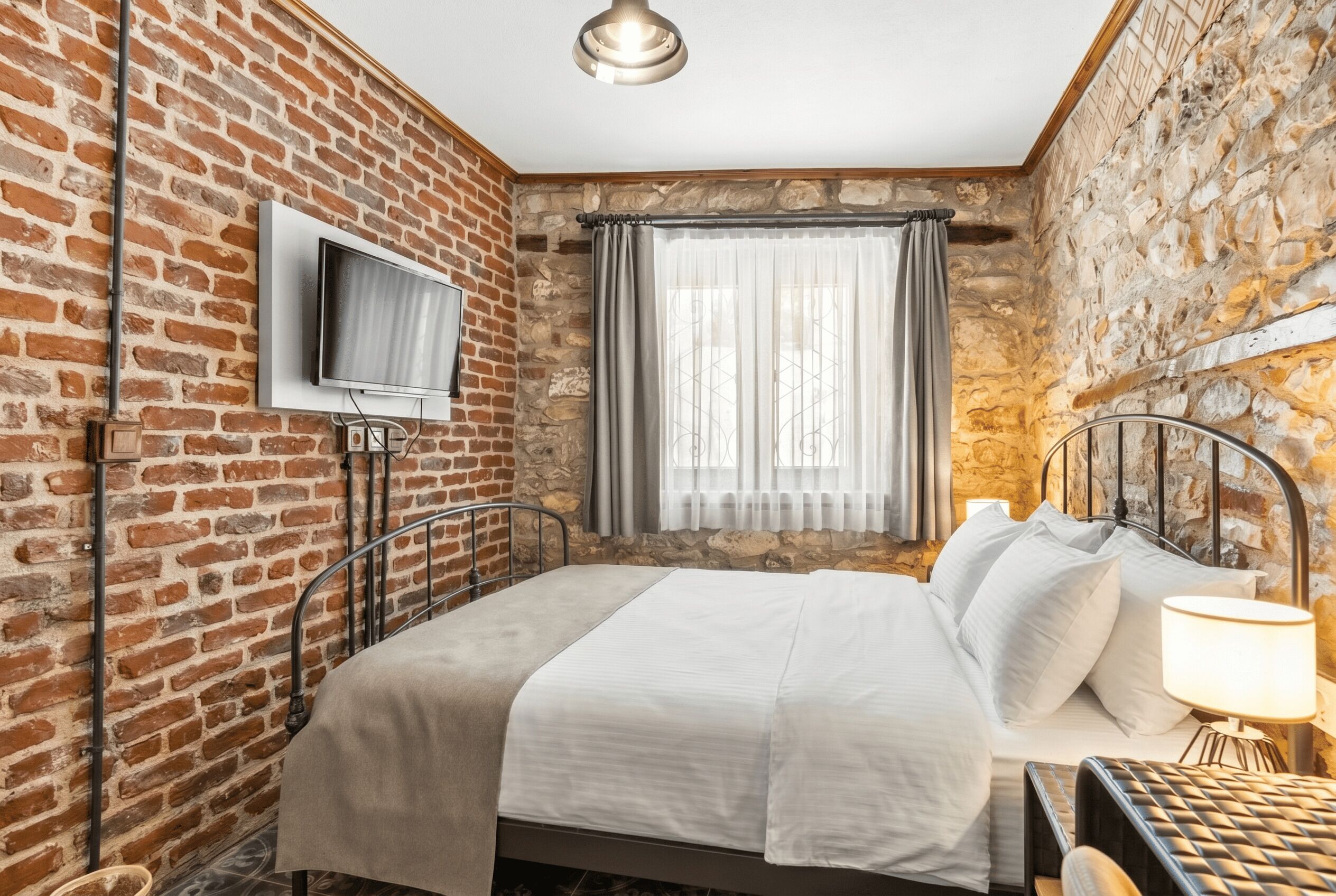 A room with a brick wall, a bed with white bedding, a television mounted on the wall, and a window with curtains.