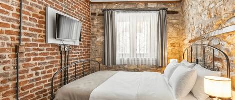 A room with a brick wall, a bed with white bedding, a television mounted on the wall, and a window with curtains.