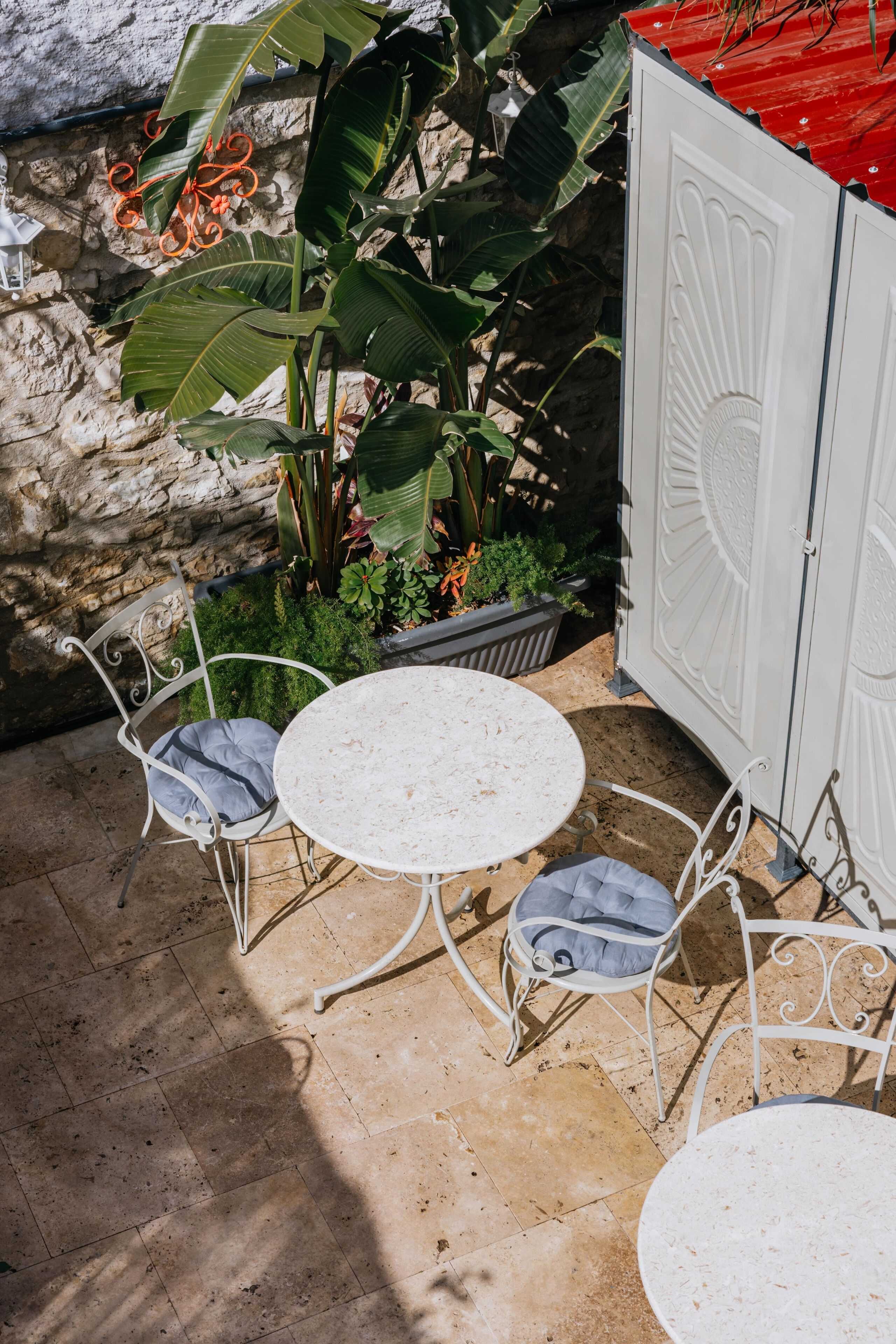 An outdoor patio with a round table and two chairs, surrounded by lush plants and a stone wall.
