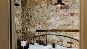 Una cama bien hecha con sábanas blancas, cabecera de metal y pared de piedra de fondo.