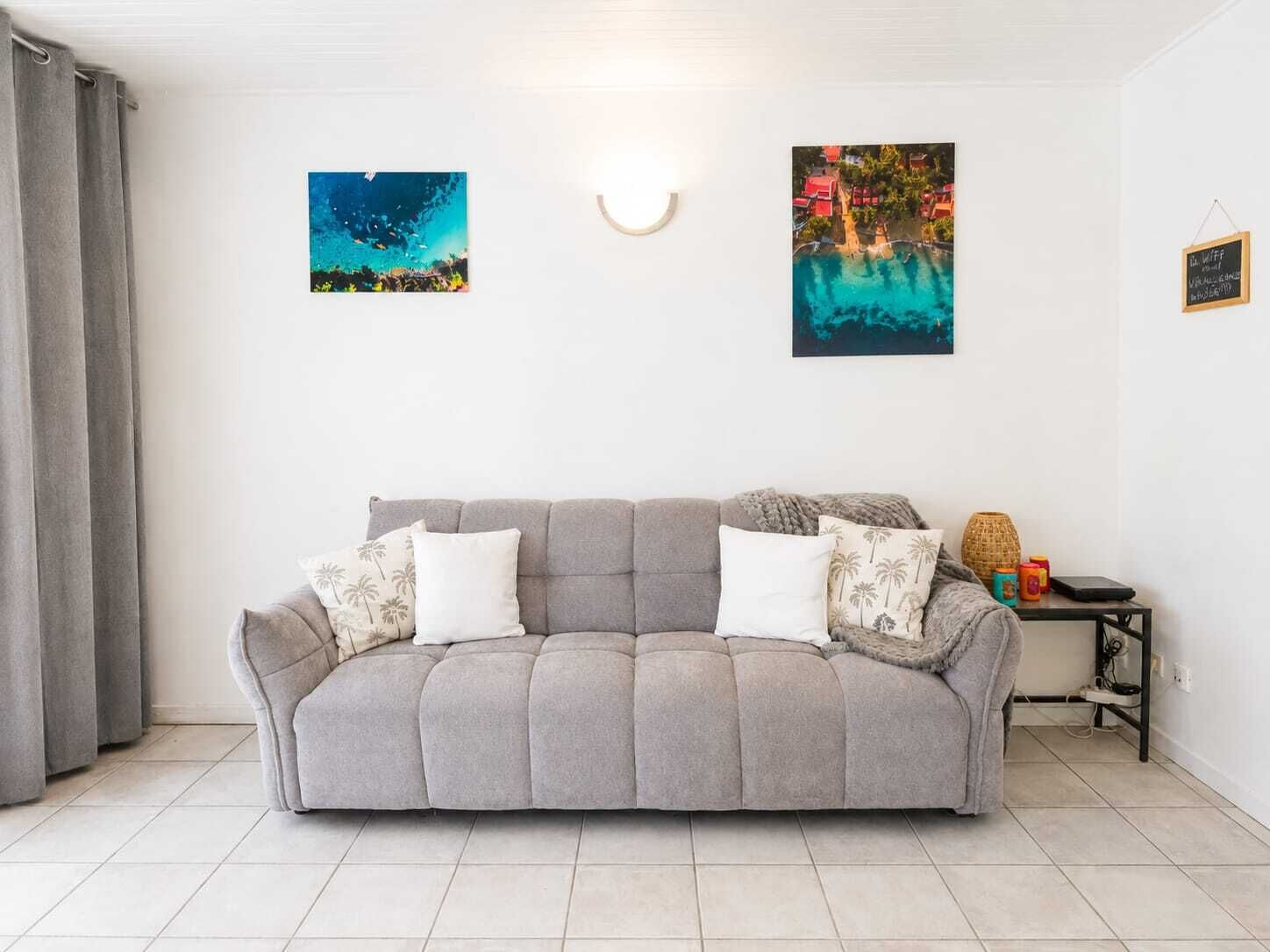 A neutral-toned living room with a grey sofa, two cushions, a small side table with a lamp, and two wall-mounted artworks.