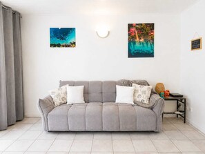 A neutral-toned living room with a grey sofa, two cushions, a small side table with a lamp, and two wall-mounted artworks. - LILI Apartment with a pool, 400 meters from the beach. (Les Trois-Îlets)