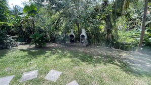 A garden with a stone pathway leading to a central area with two stone structures, surrounded by lush greenery. - LILI Apartment with swimming pool, 400m from the beach. (Les Trois-Îlets)